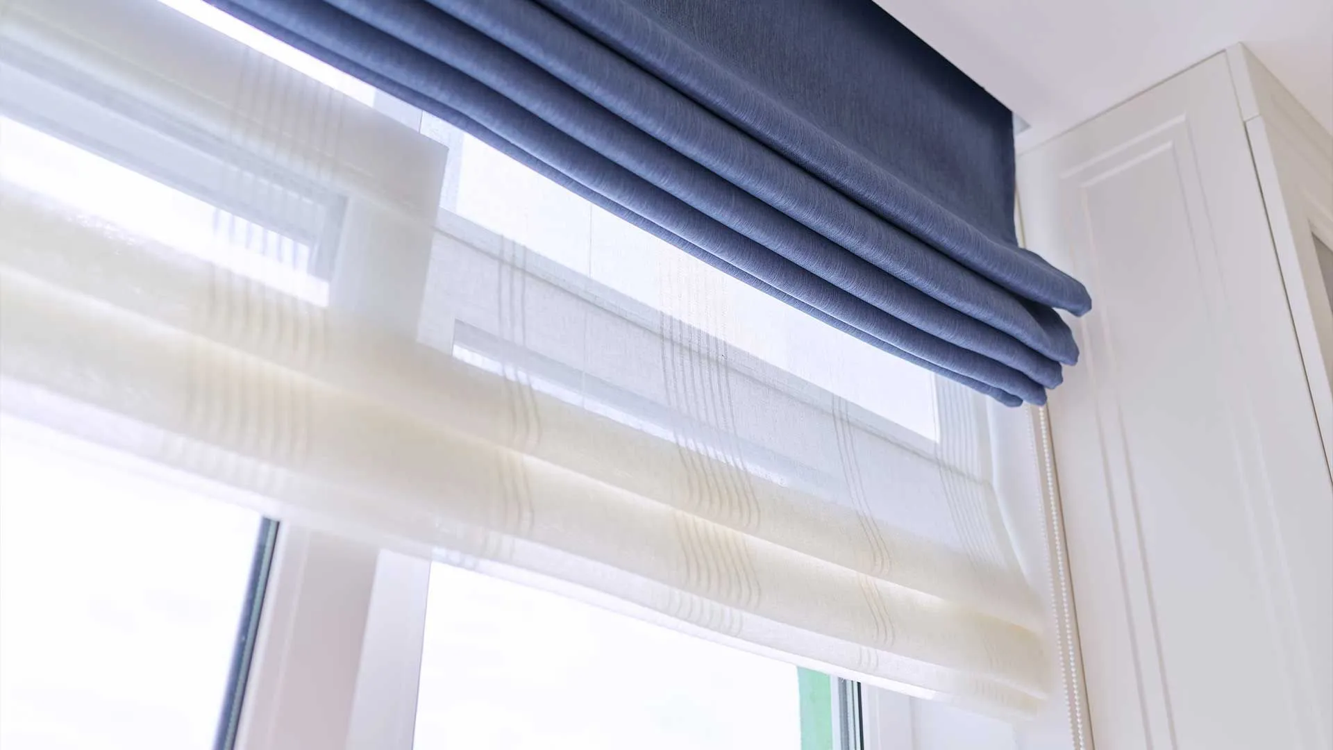 Layered blue and sheer Roman shades folded at a window